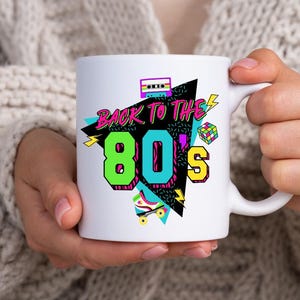 May include: White ceramic mug with a colorful graphic design featuring the text "Back to the 80's" with a cassette tape, roller skate, and geometric shapes.