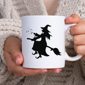 K&ouml;nnte beinhalten: Wei&szlig;e Keramiktasse mit einer schwarzen Silhouette einer Hexe auf einem Besen, Sterne von ihrem Zauberstab. Die Tasse hat einen Standardgriff und wird von einer Person gehalten, die einen cremefarbenen Pullover tr&auml;gt. Ein Halloween-Design.