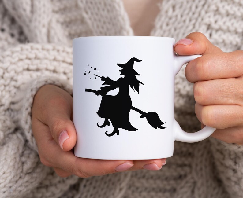 K&ouml;nnte beinhalten: Wei&szlig;e Keramiktasse mit einer schwarzen Silhouette einer Hexe auf einem Besen, Sterne von ihrem Zauberstab. Die Tasse hat einen Standardgriff und wird von einer Person gehalten, die einen cremefarbenen Pullover tr&auml;gt. Ein Halloween-Design.