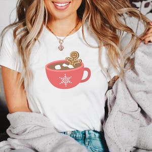 May include: White t-shirt with a red mug and a gingerbread man inside. The mug has marshmallows and a snowflake design.
