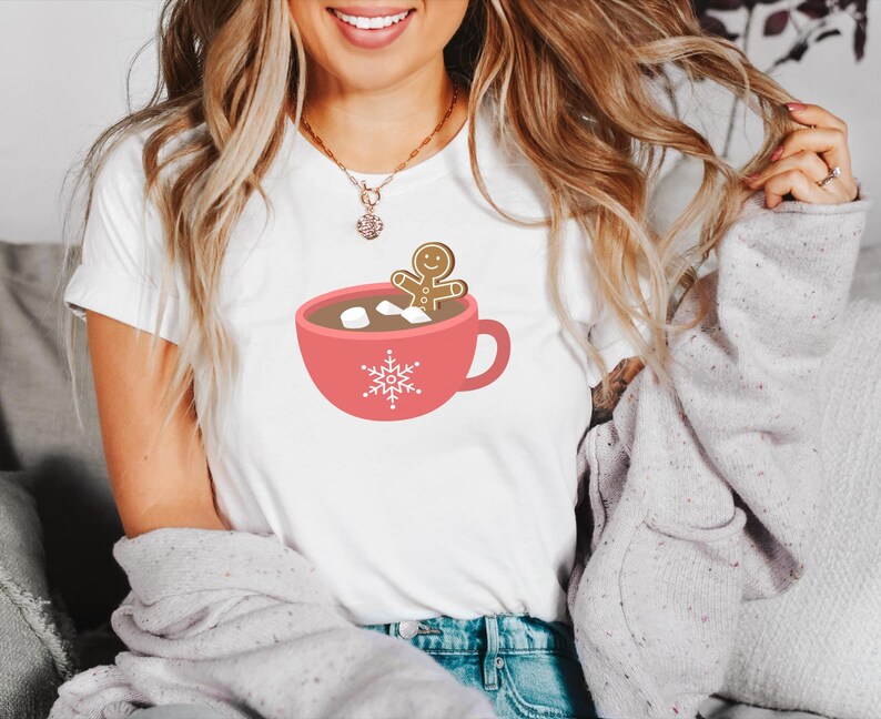 May include: White t-shirt with a red mug and a gingerbread man inside. The mug has marshmallows and a snowflake design.