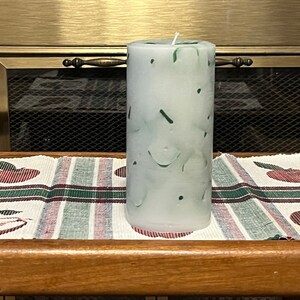 May include: A white pillar candle with green swirls and speckles. The candle is sitting on a striped table runner with red apples.