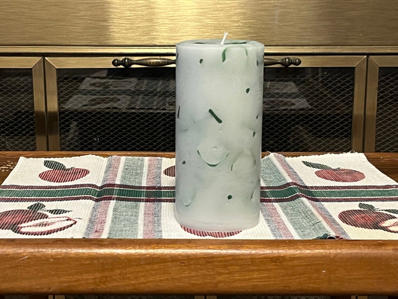 May include: A white pillar candle with green swirls and speckles. The candle is sitting on a striped table runner with red apples.