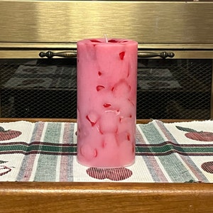 May include: A pink pillar candle with red flecks throughout. The candle is sitting on a white and green plaid table runner with red apple designs.