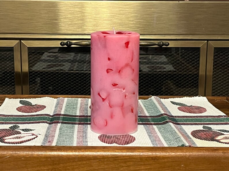 May include: A pink pillar candle with red flecks throughout. The candle is sitting on a white and green plaid table runner with red apple designs.