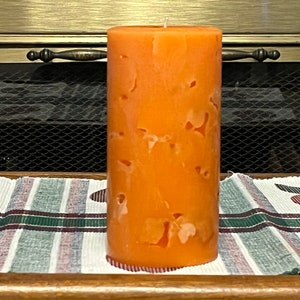 May include: A large orange pillar candle with a marbled design. The candle is sitting on a striped fabric surface.
