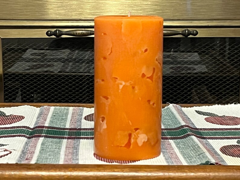 May include: A large orange pillar candle with a marbled design. The candle is sitting on a striped fabric surface.