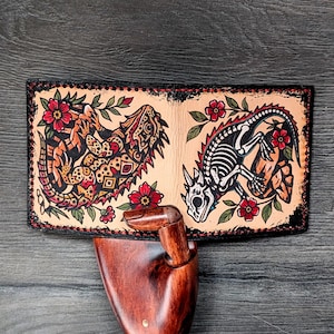 May include: A bi-fold leather wallet with a detailed tattoo-style design. One side features a colorful lizard, the other a lizard skeleton, both surrounded by red flowers and green leaves. The wallet has a black border.