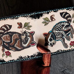 Könnte beinhalten: Eine rechteckige, cremefarbene Geldbörse mit zwei Waschbär-Illustrationen. Einer ist farbig, der andere als Skelett dargestellt. Das Design umfasst Blätter, Eicheln und Blumen mit einem schwarzen Rand.