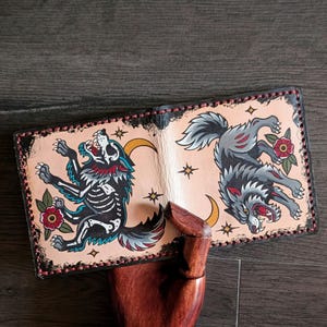 May include: Open leather wallet featuring a traditional tattoo design of two wolves. One wolf is a skeleton, the other is gray. The design includes red roses, stars, and crescent moons. The wallet is hand-stitched with a black border.