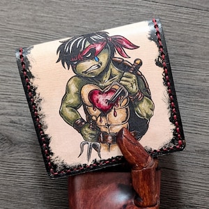 May include: A handmade leather wallet featuring a distressed image of a Teenage Mutant Ninja Turtle with a heart pierced by a dagger. The wallet has a black border with red stitching and a natural leather background.