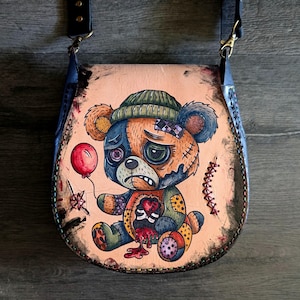 May include: A tan leather crossbody bag featuring a hand-painted, distressed teddy bear design. The bear has a sad expression, a green beanie, and a red balloon. The bag has a black strap and a dark brown border.