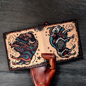 May include: A tan leather wallet featuring two stylized ram illustrations. One ram is fully colored with brown, red, and blue, while the other is a skeleton with blue and red accents. The wallet has a black border with red stitching.