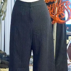May include: Black pinstripe wide-leg pants displayed on a mannequin. The pants have a high waist and a classic pinstripe pattern. The mannequin is on a pink surface, with holiday decorations in the background.