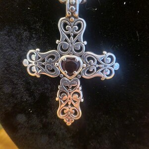 May include: Silver-toned pendant necklace featuring an ornate cross design. The cross has intricate scrollwork and a dark red gemstone at the center. The chain is a twisted rope style, and the pendant is displayed against a dark background.