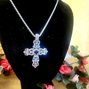 May include: A silver-toned pendant necklace featuring a detailed, openwork cross design with a dark center stone. The cross hangs from a delicate chain, displayed against a black background. The necklace is a religious or fashion accessory.