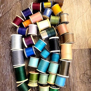 May include: A collection of assorted sewing thread spools in a wide variety of colors including blue, green, yellow, red, purple, brown, and neutral tones. The spools are arranged in a cluster with loose threads visible across the surface.