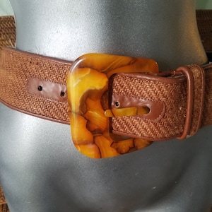 May include: A brown woven belt with a large, amber-colored plastic buckle. The buckle has a marbled pattern.