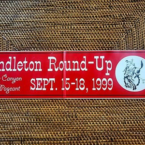May include: A red sign with white text that reads "Pendleton Round-Up" and "Happy Canyon Night Pageant." The sign also includes the date "SEPT. 15-18, 1999" and a circular graphic of a rodeo scene.