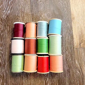 May include: A collection of twelve sewing thread spools arranged in a three-by-four grid. The spools feature a variety of solid colors including deep red, peach, light blue, white, rust, green, mint, pale green, tan, and bright red.
