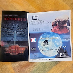 May include: Two movie-themed items: an "Independence Day" movie poster and a "Limited Edition" E.T. check. The E.T. check features the movie's logo, a blue planet, and the text "Five and 00/100 Dollars."
