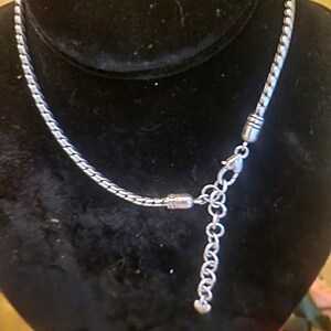 May include: A silver-toned necklace with a textured, woven chain design. The necklace is displayed on a black velvet bust, showcasing its intricate details and adjustable chain length. The clasp and extender chain are also silver.