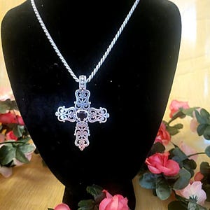 May include: A silver-colored cross pendant with an ornate, filigree design hangs from a matching chain. The cross is displayed on a black velvet bust, surrounded by pink and red roses.