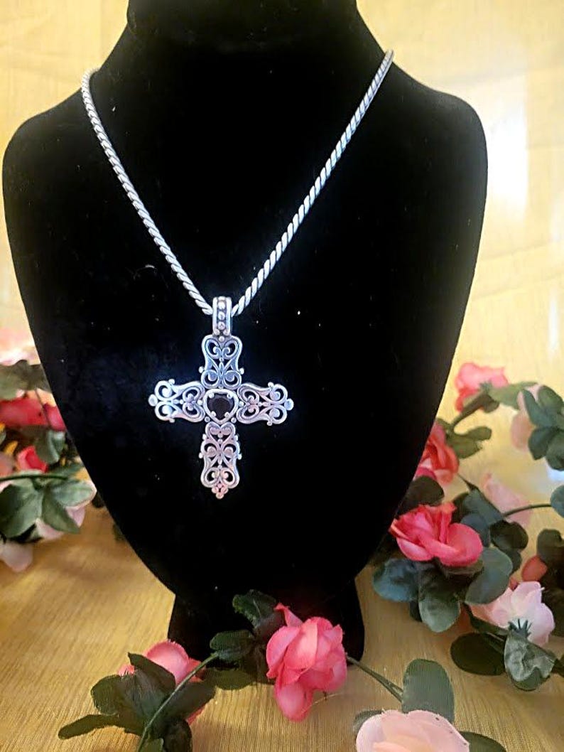 May include: A silver-colored cross pendant with an ornate, filigree design hangs from a matching chain. The cross is displayed on a black velvet bust, surrounded by pink and red roses.
