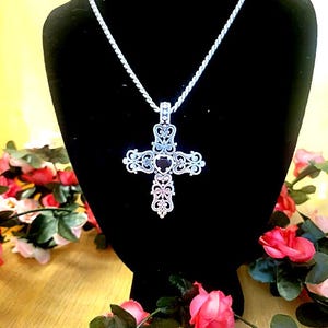 May include: Silver-toned filigree cross pendant necklace. The ornate cross design features intricate openwork patterns and is suspended from a delicate chain. The necklace is displayed on a black mannequin, surrounded by pink and red roses.