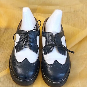 May include: A pair of black and white leather oxford shoes with a wingtip design. The shoes have black laces and a thick, black sole. The white sections are on the sides and the top of the shoe.