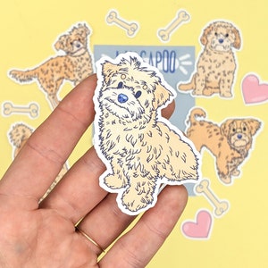May include: A sticker of a light brown fluffy dog with blue eyes. The sticker is on a hand and there are other stickers in the background, including a heart, bones, and other dogs.