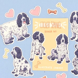 May include: A sticker set featuring Cocker Spaniel illustrations in black and white, along with pink hearts and bone-shaped stickers. The set includes a card that reads "COCKER SPANIEL STICKER SET" and "ILLUSTRATE CHRISSY" on a light yellow background, all on a light blue surface.