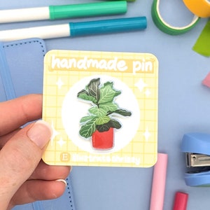 May include: A yellow and white card with the text "handmade pin" and a cartoon illustration of a potted plant with green leaves. The plant is in a red pot with a white rim.