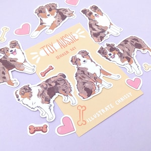 May include: A set of stickers featuring adorable cartoon Australian Shepherd dogs in various poses. The stickers are on a yellow background with the text "Toy Aussie Sticker Set" and "Illustrate Chrissy".