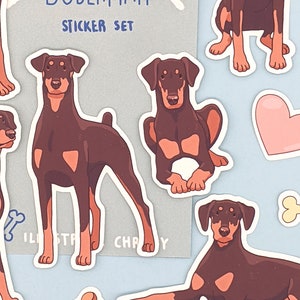May include: A sticker set featuring illustrations of Doberman Pinschers in various poses. The stickers are on a light blue background with a bone and a heart. The text "Doberman Sticker Set" is on a gray background.