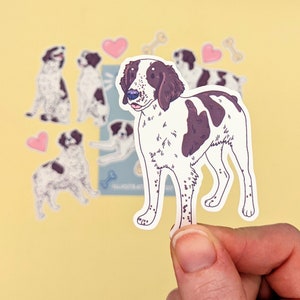 May include: A sticker of a brown and white dog with black spots. The dog is standing on all four legs and has its tongue sticking out. The sticker is on a yellow background.