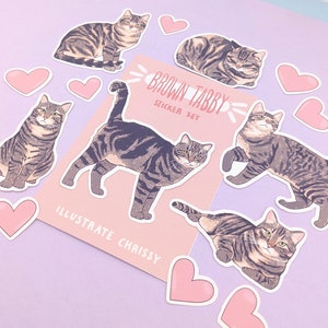 May include: A set of brown tabby cat stickers with pink hearts. The stickers feature different poses of the cat, including sitting, lying down, and walking. The stickers are labelled "Brown Tabby Sticker Set" and "Illustrate Chrissy".