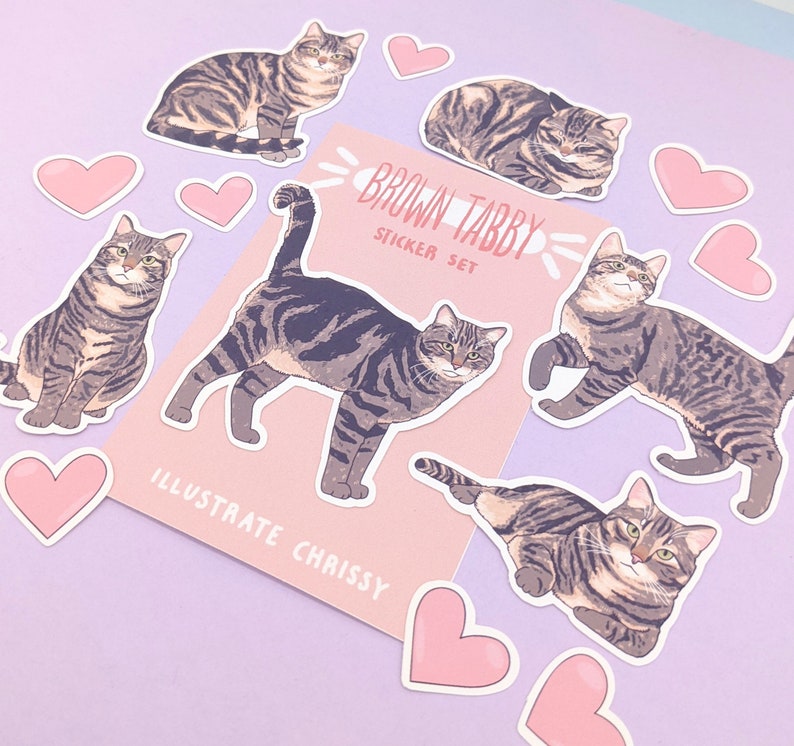 May include: A set of brown tabby cat stickers with pink hearts. The stickers feature different poses of the cat, including sitting, lying down, and walking. The stickers are labelled "Brown Tabby Sticker Set" and "Illustrate Chrissy".