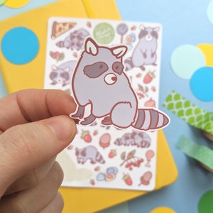 May include: A gray raccoon sticker with a brown outline and a striped tail is held in front of a sheet of raccoon-themed stickers. The sheet includes various raccoon illustrations, strawberries, and sweets. A yellow notebook and colorful paper circles are in the background.