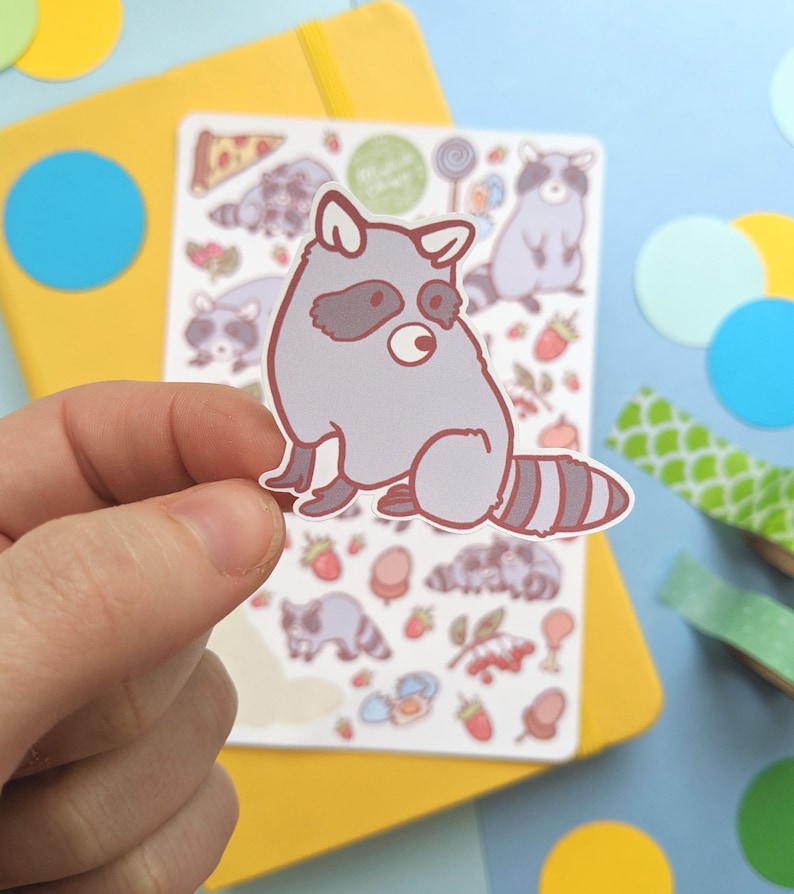 May include: A gray raccoon sticker with a brown outline and a striped tail is held in front of a sheet of raccoon-themed stickers. The sheet includes various raccoon illustrations, strawberries, and sweets. A yellow notebook and colorful paper circles are in the background.