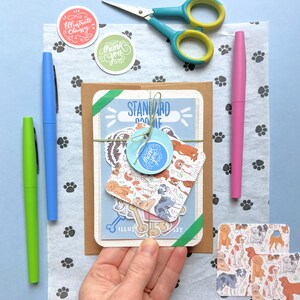 Pu&ograve; includere: Una busta marrone con un biglietto blu e bianco all'interno. Il biglietto ha un'illustrazione di un cane e il testo "Standard Doodle". Ci sono diversi adesivi sul biglietto, tra cui uno che dice "Grazie".