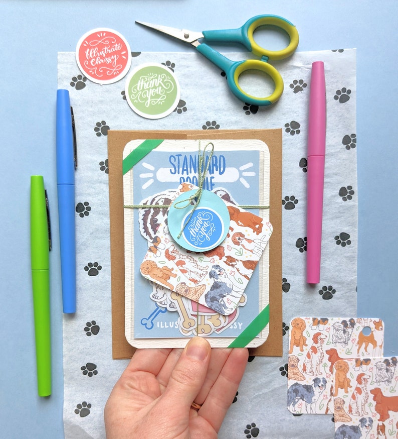 Pu&ograve; includere: Una busta marrone con un biglietto blu e bianco all'interno. Il biglietto ha un'illustrazione di un cane e il testo "Standard Doodle". Ci sono diversi adesivi sul biglietto, tra cui uno che dice "Grazie".
