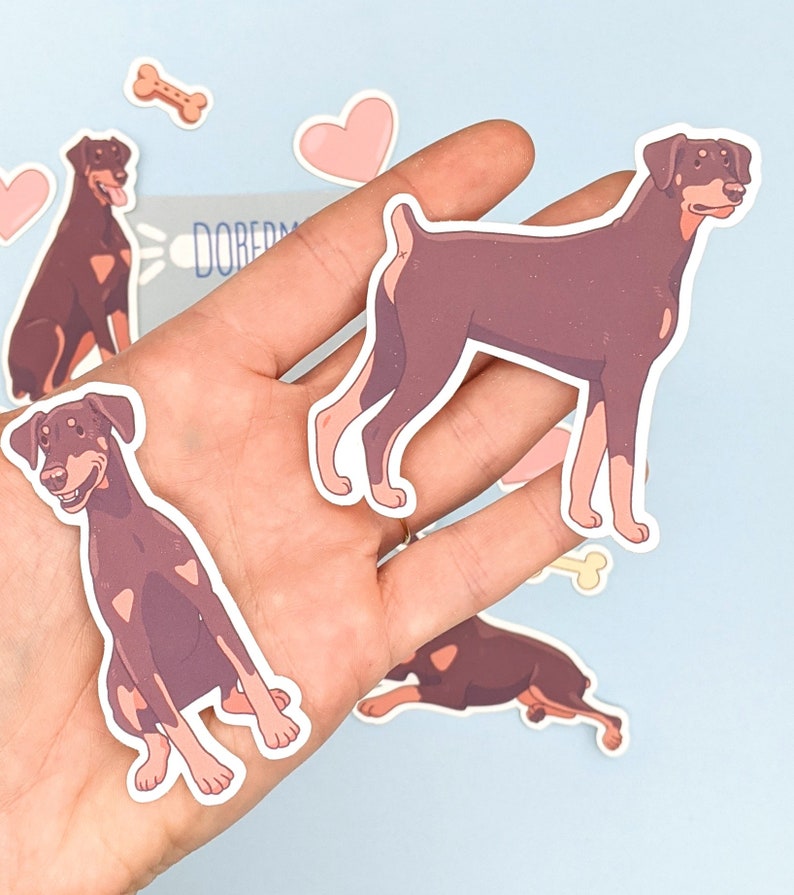 May include: A set of five stickers featuring brown and black Doberman Pinscher dogs in various poses. The stickers are on a light blue background with pink hearts and a bone.