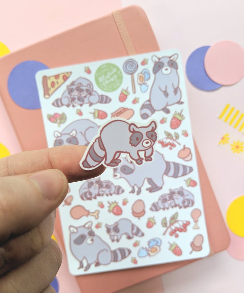 May include: A sheet of raccoon-themed stickers. The stickers feature various raccoon illustrations with food items like pizza, strawberries, and candy. The stickers are in shades of gray, brown, and pink, with a light blue background.