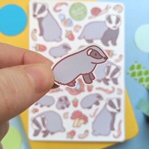 K&ouml;nnte beinhalten: Eine Bogen mit Aufklebern, die eine Vielzahl von niedlichen Cartoon-Dachsen in verschiedenen Posen zeigen. Die Aufkleber sind auf wei&szlig;em Papier gedruckt und haben eine gl&auml;nzende Oberfl&auml;che. Die Dachse sind alle helllila mit braunen Akzenten. Auf dem Bogen befinden sich auch eine Vielzahl anderer Aufkleber, darunter Pilze, Erdbeeren und W&uuml;rmer.