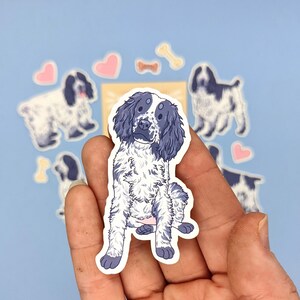 May include: A sticker featuring a seated dog with a blue and white coat. The dog has floppy ears and a friendly expression. Surrounding the dog are other dog stickers, hearts, and bone shapes, all on a light blue background.