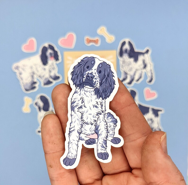May include: A sticker featuring a seated dog with a blue and white coat. The dog has floppy ears and a friendly expression. Surrounding the dog are other dog stickers, hearts, and bone shapes, all on a light blue background.