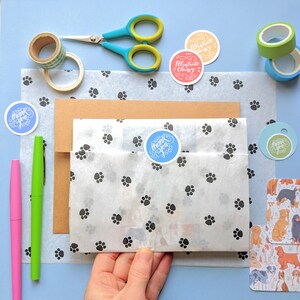 K&ouml;nnte beinhalten: Eine wei&szlig;e Geschenkt&uuml;te mit Pfotenabdruckmuster ist mit braunem Papier und einem blauen Dankesch&ouml;n-Aufkleber verpackt. Die Tasche ist umgeben von verschiedenen Bastelartikeln, darunter eine Schere, Washi-Tape und Aufkleber mit dem Text "Illustrate Classy".