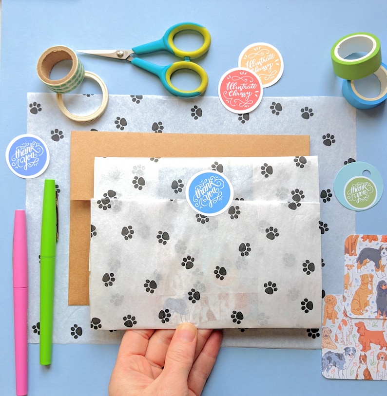 K&ouml;nnte beinhalten: Eine wei&szlig;e Geschenkt&uuml;te mit Pfotenabdruckmuster ist mit braunem Papier und einem blauen Dankesch&ouml;n-Aufkleber verpackt. Die Tasche ist umgeben von verschiedenen Bastelartikeln, darunter eine Schere, Washi-Tape und Aufkleber mit dem Text "Illustrate Classy".
