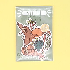 May include: A sticker set featuring cute cartoon sloths in a jungle setting. The set includes a sloth hanging from a branch, a sloth sitting on a branch, and a leaf. The stickers are labeled "Sloth Sticker Set" and "Smooth Matte".